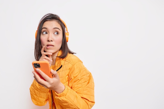 Задумчивая азиатская женщина-модель кусает губы с задумчивым взглядом, использует смартфон и стереонаушники для прослушивания музыки, одетая в куртку, изолированную на белой стене с пустым пространством справа