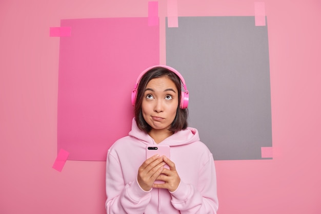 Задумчивая азиатская девушка любит новое приложение на смартфоне для песен плейлистов, слушает любимую музыку, сосредоточенную выше, задумчиво, одетая в толстовку, позирует на розовой стене, думает о чем-л.