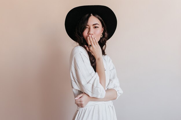 Pensive asian woman in black hat looking at camera. Studio shot of glamorous japanese model isolated on beige background.