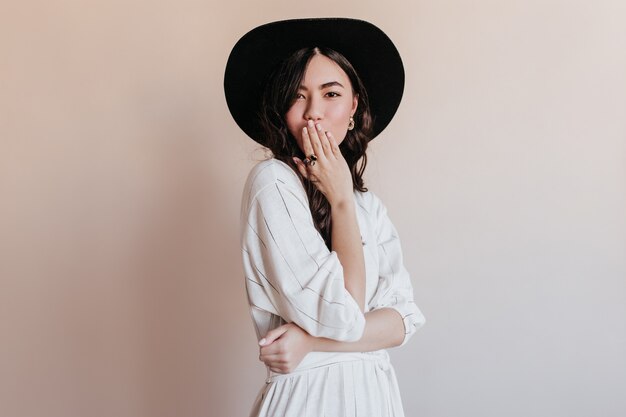 카메라보고 검은 모자에 잠겨있는 아시아 여자. 베이지 색 배경에 고립 된 매력적인 일본 모델의 스튜디오 샷.