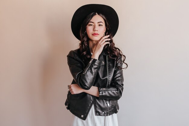 Pensive asian young woman in hat looking at camera. Japanese woman in leather jacket standing on beige background.