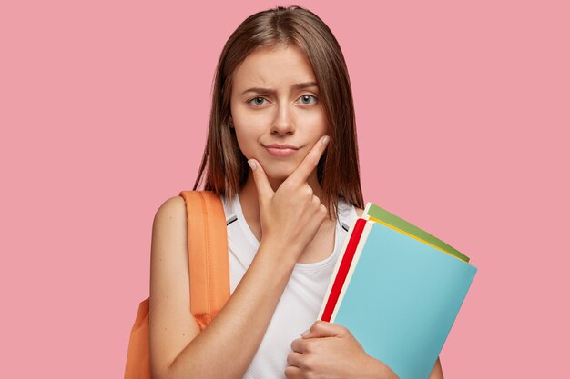 Pensive attractive Cauasian girlfriend holds chin and looks with puzzlement, has straight hair, carries rucksack