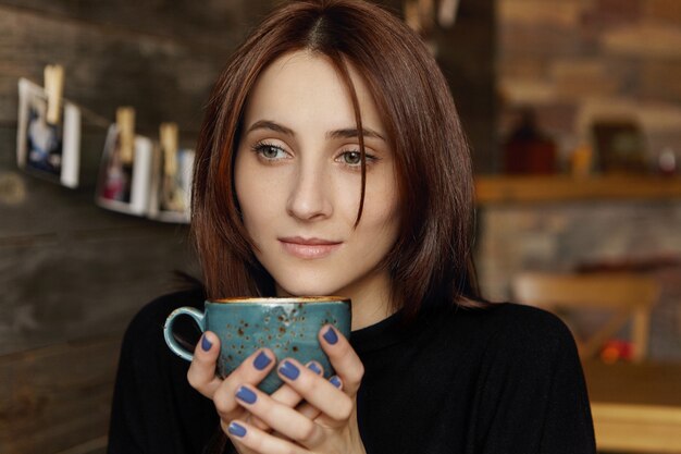 Pensive attractive young European female with brown chocolate hair wearing elegant black dress holding cup of cappuccino, daydreaming, enjoying hot and fresh drink while sitting at cozy coffee shop