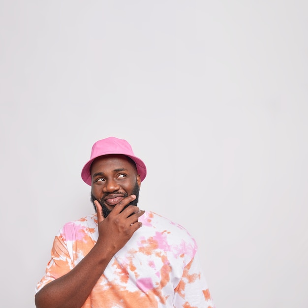 Free photo pensive bearded man holds chin looks above with tricky happy expression thinks about something wears pink panama colorful t shirt isolated over white bacground copy space above