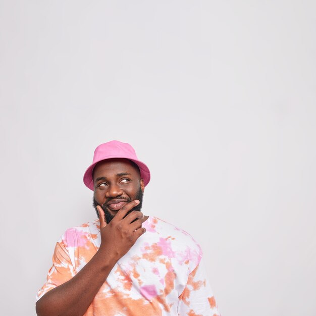 Pensive bearded man holds chin looks above with tricky happy expression thinks about something wears pink panama colorful t shirt isolated over white bacground copy space above