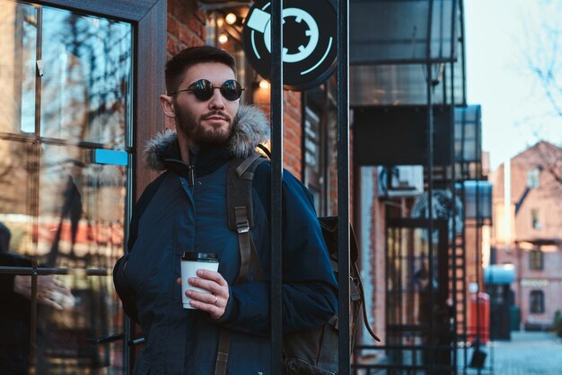 Pensive bearded man in sunglasses is drinking hot beverage while walking on the street.