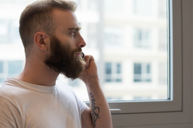 Pensive bearded man talking on phone and looking out window