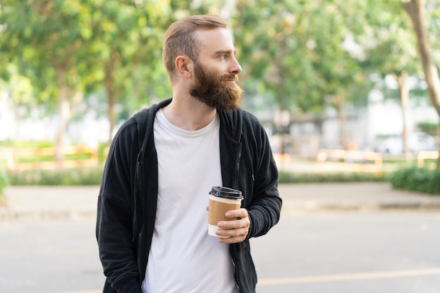 Задумчивый бородатый мужчина гуляет по городу и держит пластиковый стаканчик
