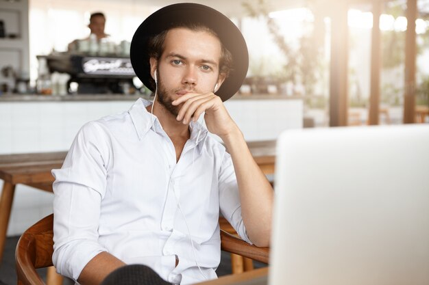 Pensive bearded student in black hat holding hand on his chin sitting in front of open laptop computer and listening to audio course on earphones while studying online during lunch at cafeteria