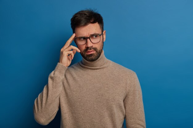 Free photo pensive bearded young man keeps finger on temple, recalls necessary information in mind, wears transparent glasses and casual jumper