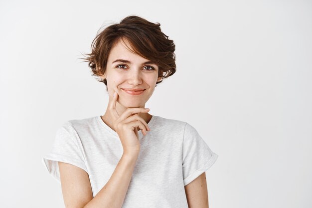 Pensive beautiful woman with short messy hair, smiling and touching clean perfect skin without makeup, standing on white wall