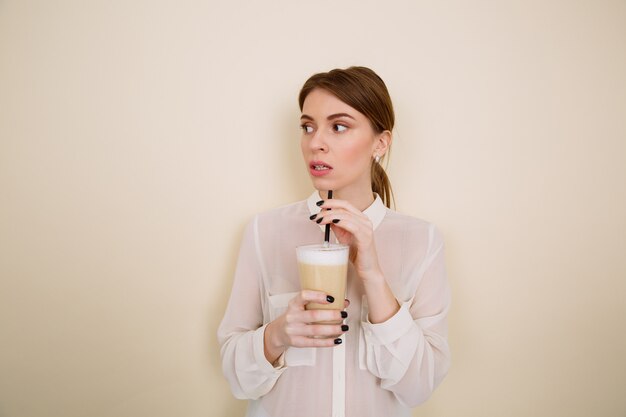 Pensive beautiful young woman standing and drinking coffee with milk