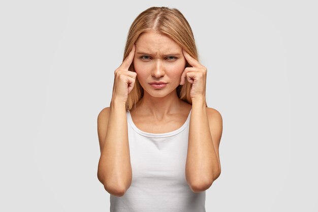 Pensive beautiful young woman with light straight hair, keeps fingers on temples, feels pain