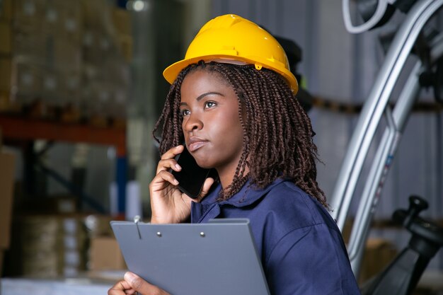 창고에 서서 핸드폰에 말하는 hardhat 잠겨있는 흑인 여성 엔지니어. 배경에 상품이있는 선반. 공간을 복사하십시오. 노동 또는 커뮤니케이션 개념
