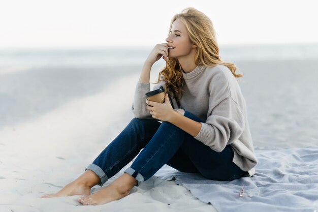 Pensive blinde woman in jeans sitting in sand and looking at sea. Outdoor portrait of relaxed caucasian woman drinking coffee at beach.