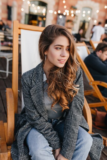 Pensive blue-eyed girl with pale skin sitting in recliner in outdoor cafe and looking away