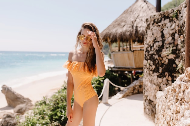 Pensive brown-haired girl in orange swimsuit posing in front of bungalow. Outdoor shot of pretty white young lady in sunglasses chilling at ocean resort.