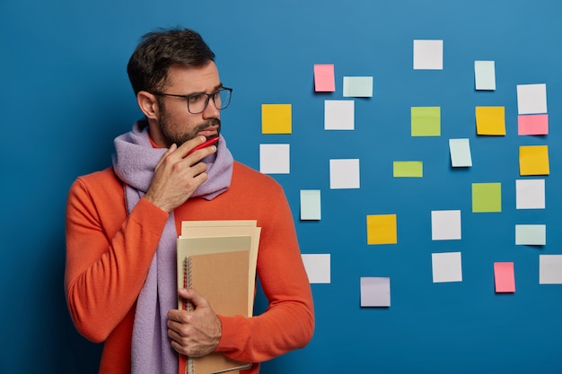 Free photo pensive brunet male student holds chin, has thick bristle, holds necessary notepads, wears scarf and sweater