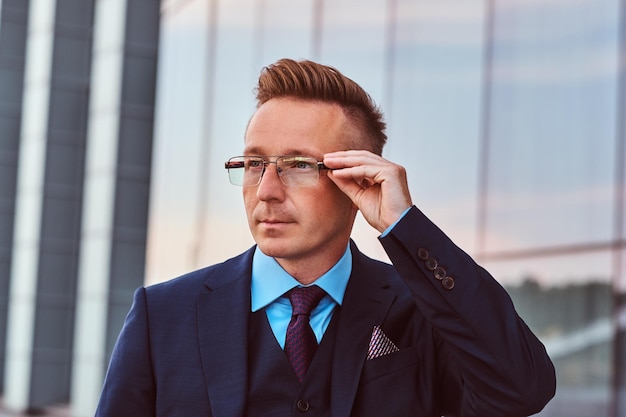 Задумчивый бизнесмен, одетый в элегантный костюм, смотрит в сторону и поправляет очки, стоя на улице на фоне небоскреба.