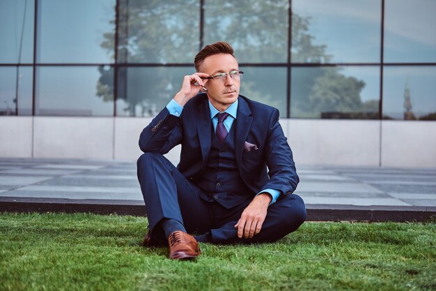 Pensive businessman dressed in an elegant suit sitting on a green lawn against cityscape background.