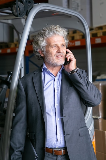 Free photo pensive businessman standing in warehouse near forklift and talking on cellphone