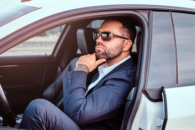 Pensive businessman with tattoo on his arm in sunglasses dressed in a formal suit sitting on the front seats in the luxury car.