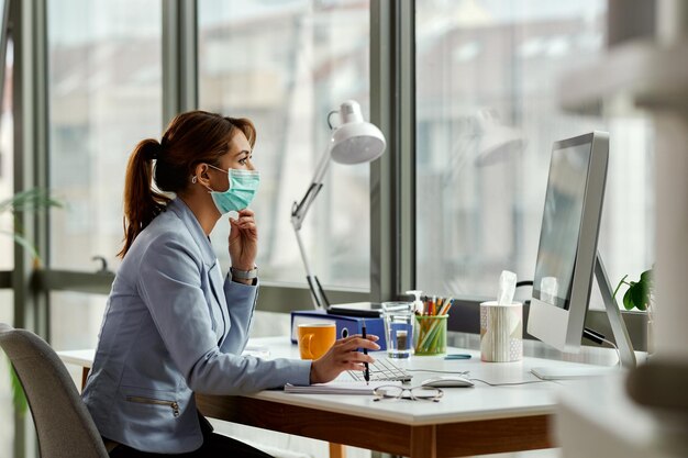 Задумчивая деловая женщина в маске для лица читает электронную почту на настольном ПК в офисе