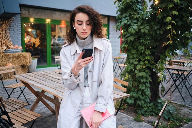 Pensive casual brunette girl in trench coat thoughtfully using cellphone on city street