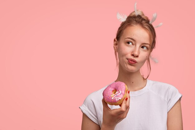 Pensive Caucasian female with feathers on head, looks thoughtfully aside, holds delicious sweet doughnut, dressed in casual white t-shirt, stands against pink wall with blank space for text