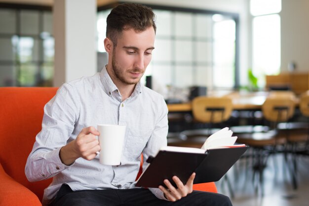 カップを持って読書をしている夢中の白人の若い男