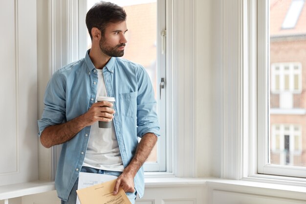 Pensive chairman with dark bristle, dressed in stylish shirt, holds takeaway coffee, papers, studies taxes