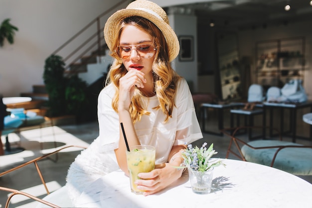 Pensive curly girl in vintage straw hat and white dress waiting boyfriend in cafe