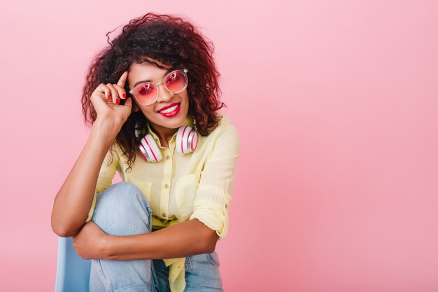 Pensive curly girl in yellow shirt embracing her knee and smiling. Indoor portrait of fashionable african woman in sunglasses sitting on chair.