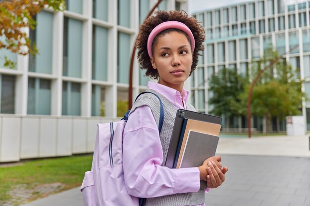 Pensive curly haired student carries notepads and digital tablet strolls at urban setting enjoys resting outdoors dressed in casual clothes carries backpack stands against modern glass building