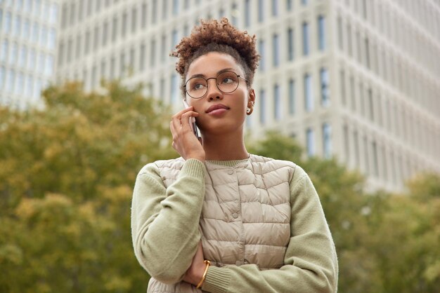 Pensive curly haired woman has telephone conversation focused into distance with thoughtful expression wears round spectacles casual clothing strolls outdoors at street against blurred background