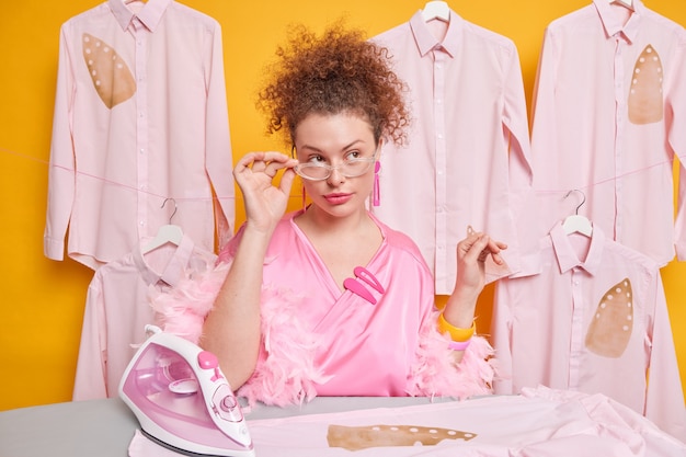 Pensive curly haired woman keeps hand on rim of spectacles dressed in silk gown poses near ironing board with electric iron poses against shirts on hangers thinks how to finish domestic work in time