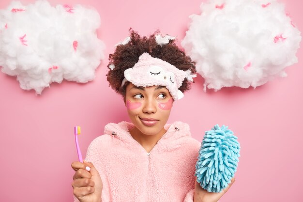 pensive curly haired woman with feathers on head undergoes beauty and hygiene procedures after awakening holds toothbrush shower sponge looks thoughtfully away isolated over pink wall.