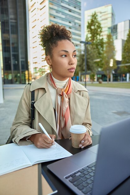 Задумчивая кудрявая женщина записывает информацию в блокнот, пьет кофе, смотрит обучающее видео через портативный компьютер, позирует в пальто на фоне городских зданий