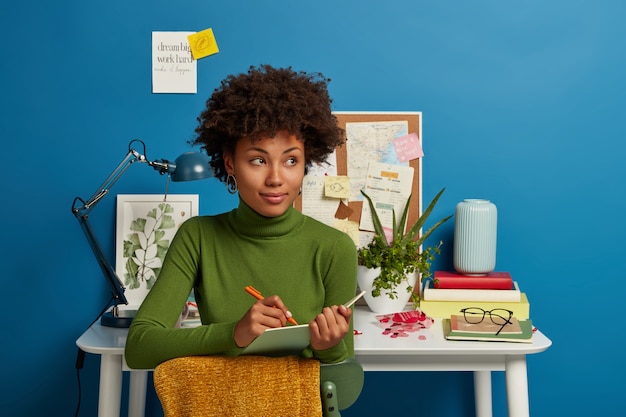 Free photo pensive curly young girl writes down future plans and goals in notepad, thinks about good idea