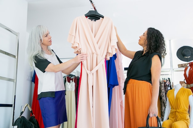 物思いにふける顧客と店の売り手が一緒にラックでドレスを閲覧し、ファッション店で服を選びます。側面図。ショッピングや小売の概念