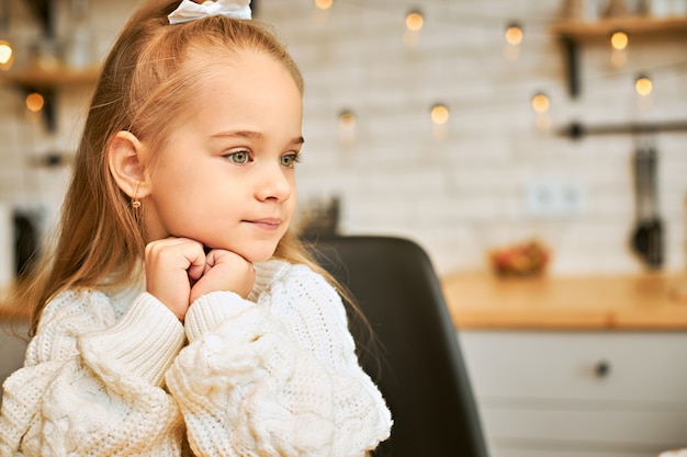 Pensive cute European little girl in knitted jumper holding both hands at her face and looking away, thinking about something, waiting mother from work. Adorable baby child sitting in kitchen alone