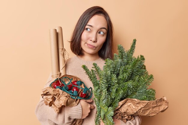 Pensive dark haired woman concentrated away looks with dreamy expression away holds spruce branches wrapped in paper retro garland prepares for winter holidays isolated over beige background