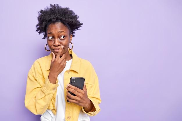 Задумчивая темнокожая девочка-подросток с причесанными кудрявыми волосами разговаривает по современному мобильному телефону, сосредоточенно вдали, с задумчивым выражением лица носит желтую куртку, изолированную на фиолетовом