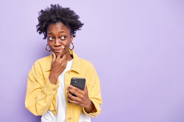 Pensive dark skinned teenage girl with combed curly hair uses modern mobile phone chatting focused away has thoughtful expression wears yellow jacket isolated on purple 