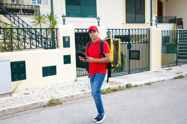 物思いにふける配達員が住所を探し、タブレットを持っています。黄色のサーモバッグを運び、注文を配達する赤い帽子とシャツのプロの宅配便。配送サービスとオンラインショッピングのコンセプト
