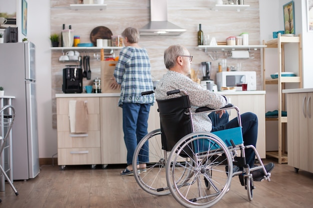 휠체어를 탄 잠겨있는 장애인 노인이 부엌에서 창문을 바라보고 있습니다. 아내가 아침 식사를 준비하는 동안 창밖을 내다보며 휠체어에 앉아 있는 장애인 남자. 유효하지 않음, p