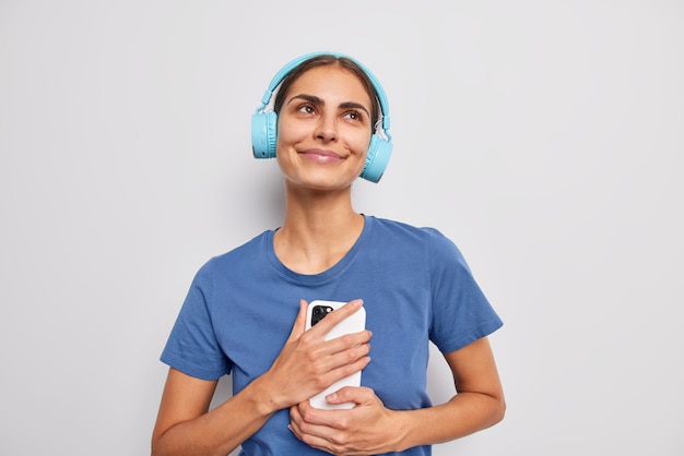 Задумчивая мечтательная брюнетка использует беспроводные наушники и смартфон слушает приятную песню, носит повседневную футболку, изолированную на белом фоне Хобби людей и концепция современных технологий