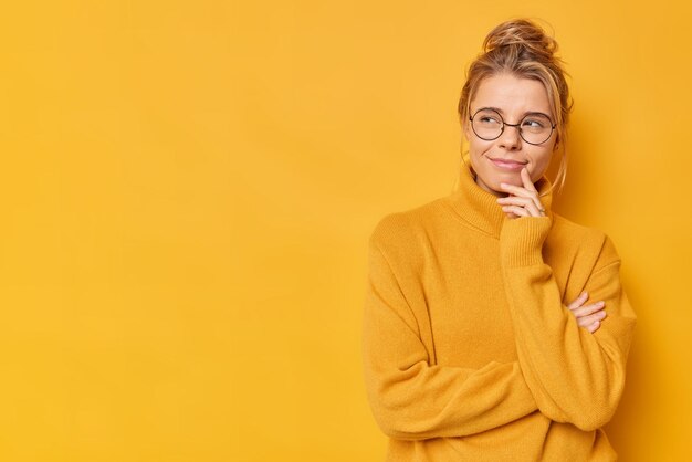 Pensive dreamy young European woman keeps hand near mouth looks suspicious away wears round spectacles and jumper isolated over yellow backgroud copy space aside for your promotional content