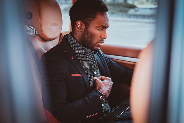Pensive elegant afro etnicity businessman is sitting in the car as a passenger while working on his laptop.