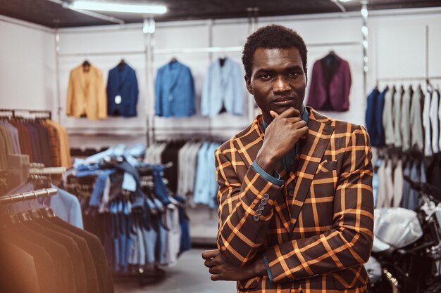 Pensive elegantly dressed African-American man posing with hand on chin while standing in a classic menswear store, looking at camera.